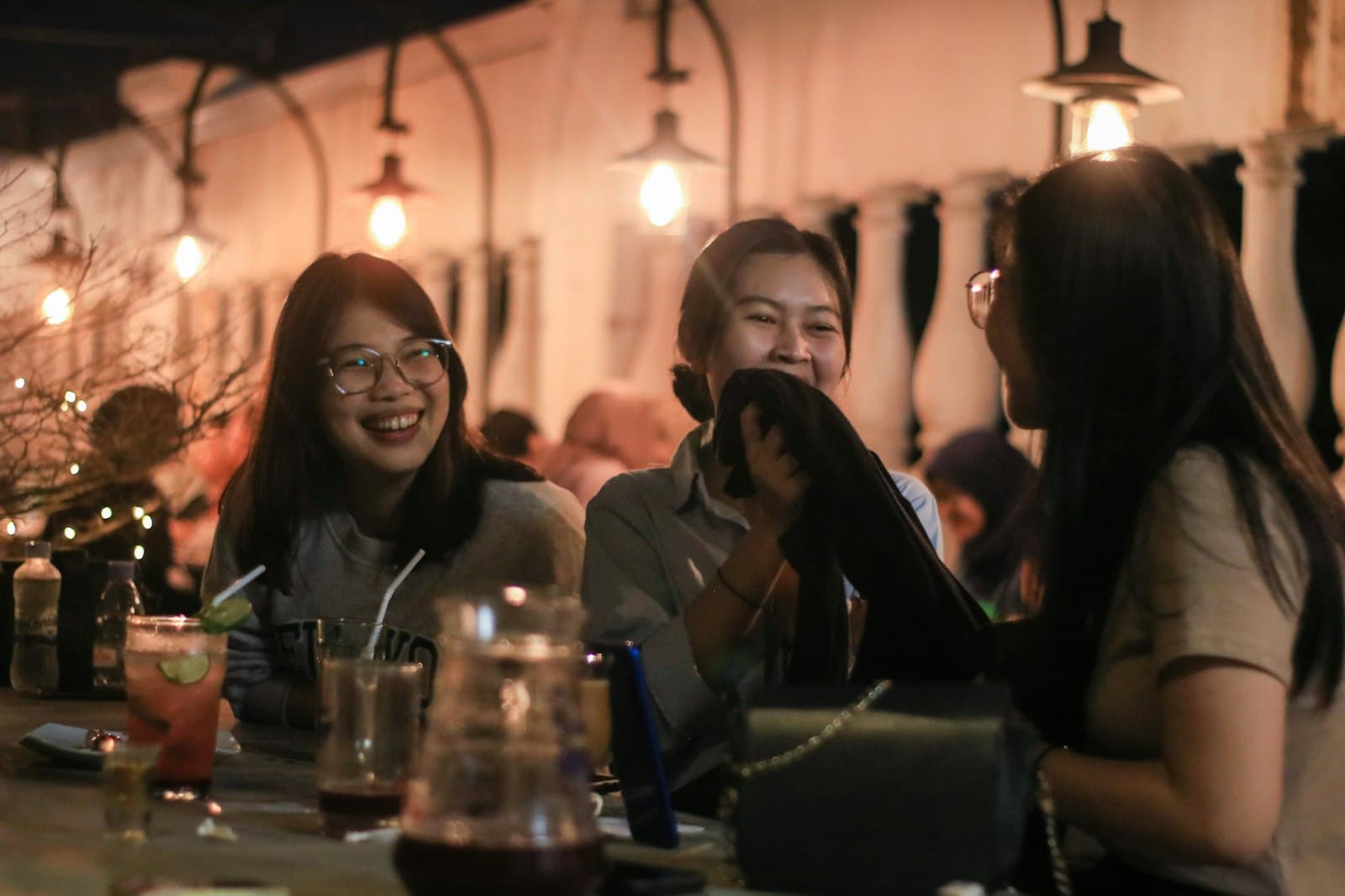 Três mulheres rindo numa mesa comprida à noite, com lâmpadas acesas em cima